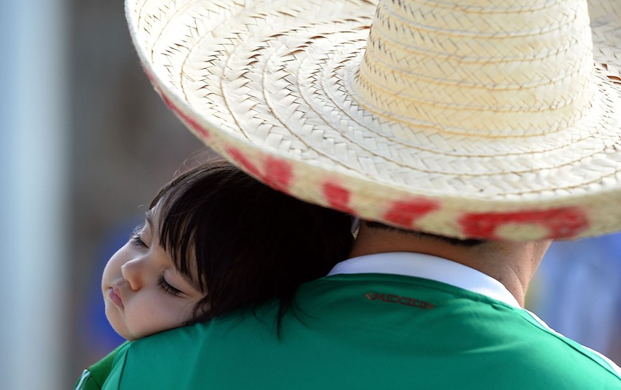 A young child sleeps while resting on the shoulder of an adult, who is wearing a sombrero.