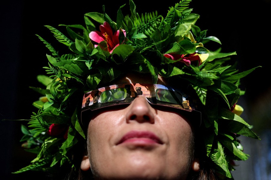 A woman wearing a wreath of leaves and mirrored eclipse glasses looks up toward the sun.