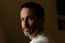 A white man wearing a white shirt stands in front of a yellow wall
