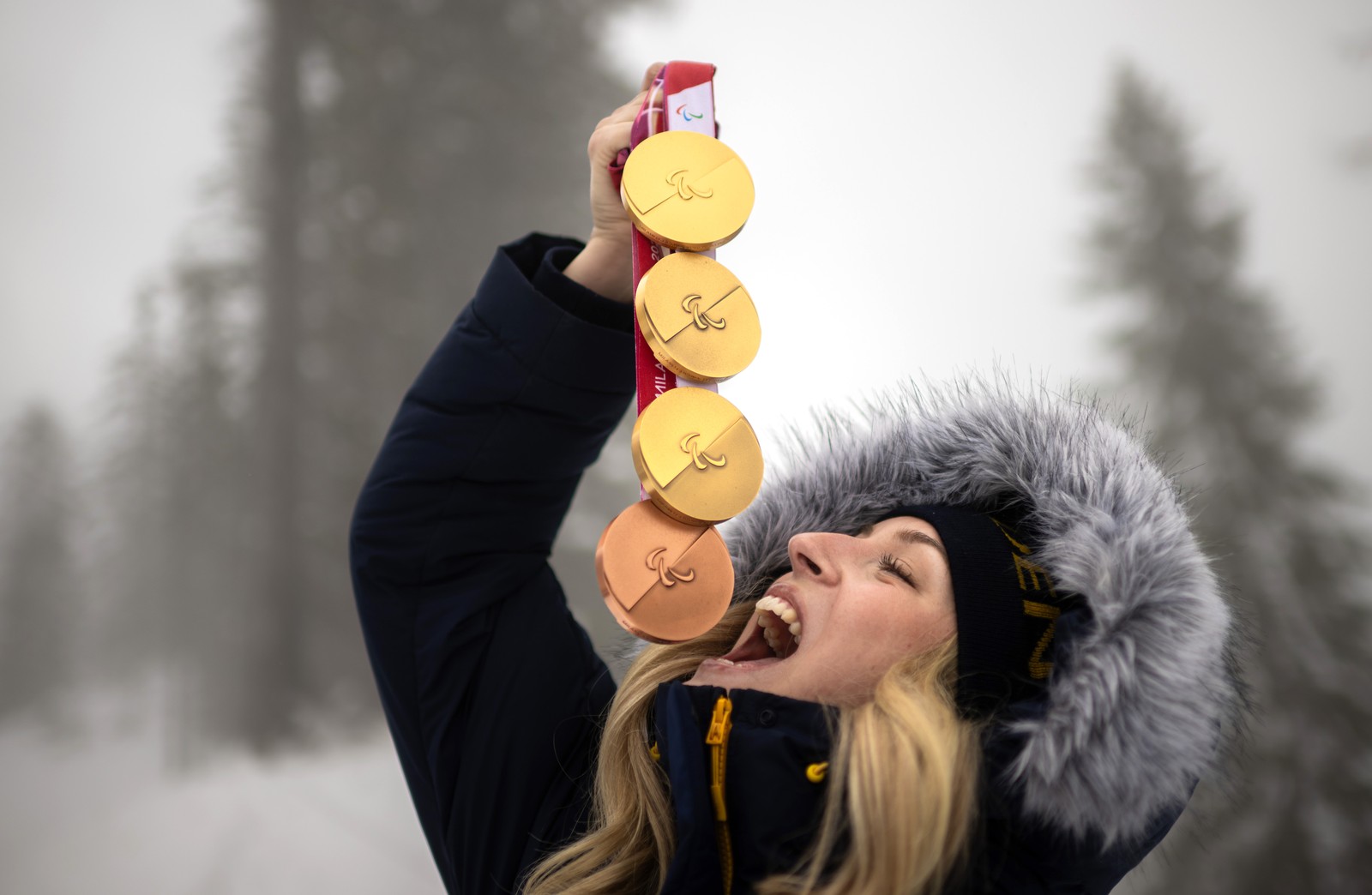 An athlete in a warm coat stands outside for a photo shoot, holding four Paralympic medals above their upturned face, pretending as if they might swallow them.