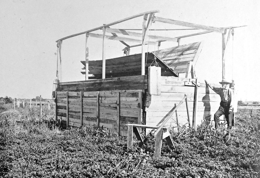 A person stands beside a large wooden paddle-wheel style windmill.