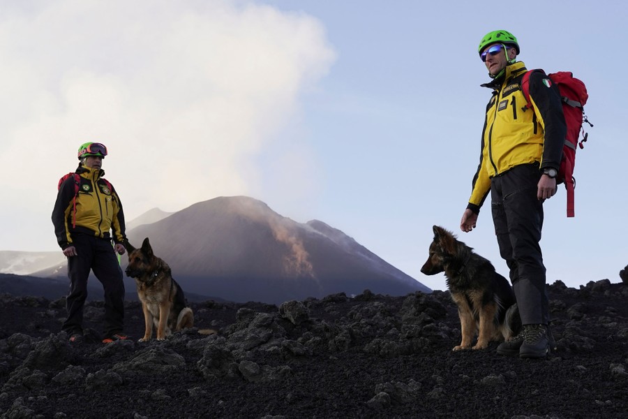 Two people, each with a German shepherd, stand on volcanic material near a volcanic mound.