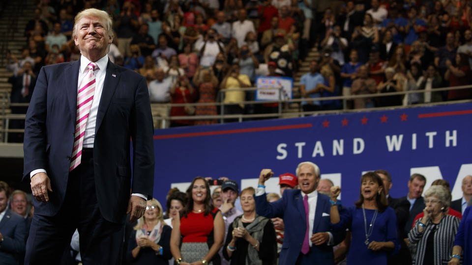 President Trump smiles at a campaign rally, with clapping supporters seen behind him.