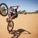 A child pops a wheelie on a bike on a patch of dirt near some houses.