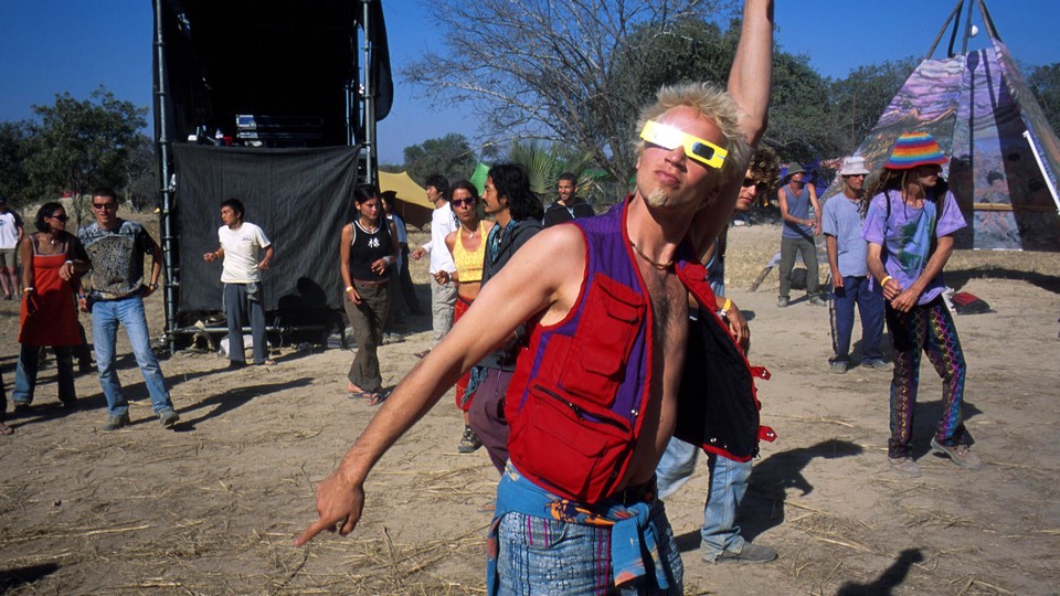 A man wearing eclipse glasses and colorful clothing gestures upward.