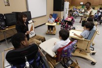 A woman holds a board with a cat drawing and the letter "C." A semicircle of students with disabilities surrounds her