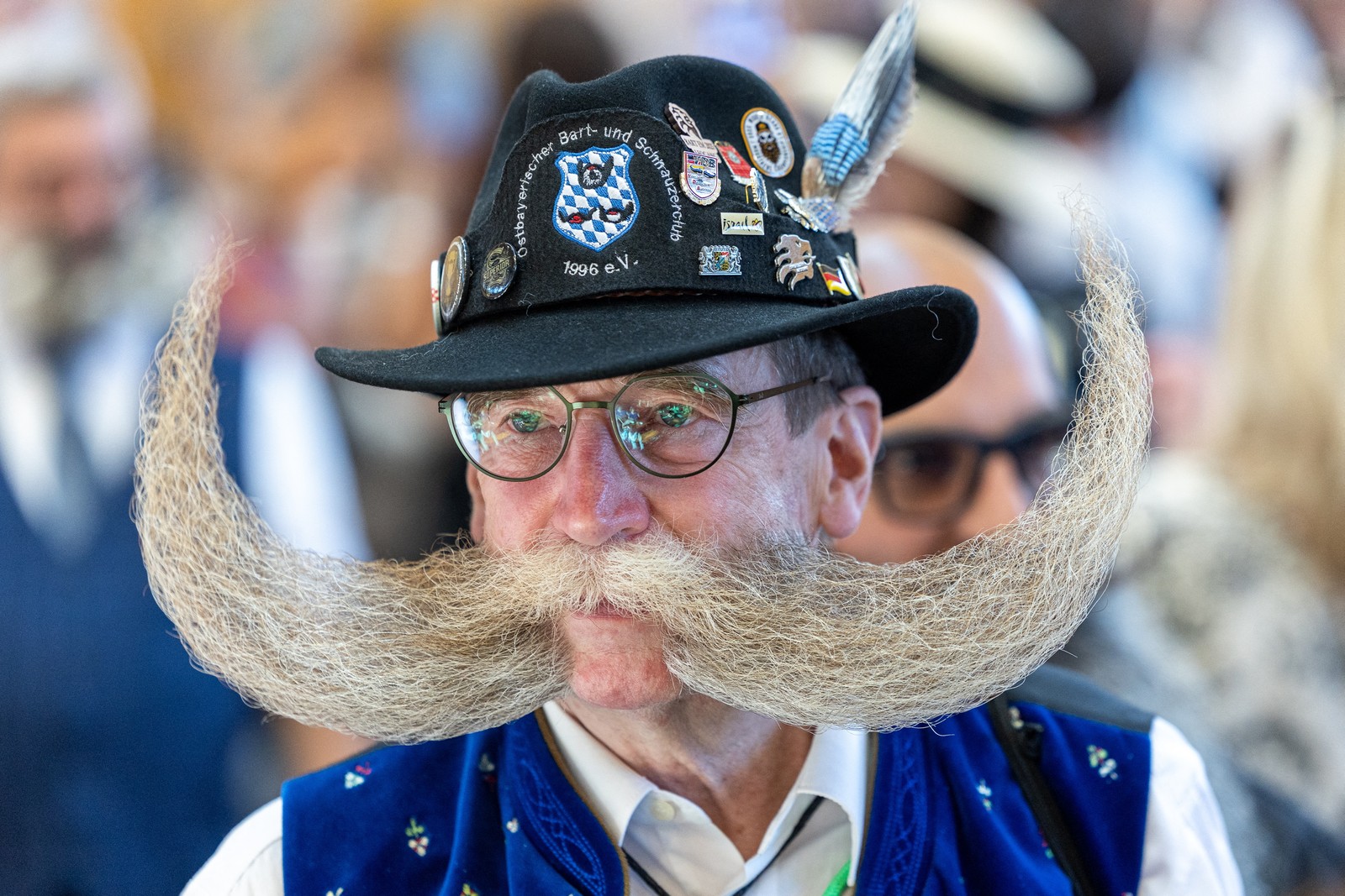 A man poses, displaying his enormous curved mustache and facial hair that stretches out to the side, beyond the brim of his hat.