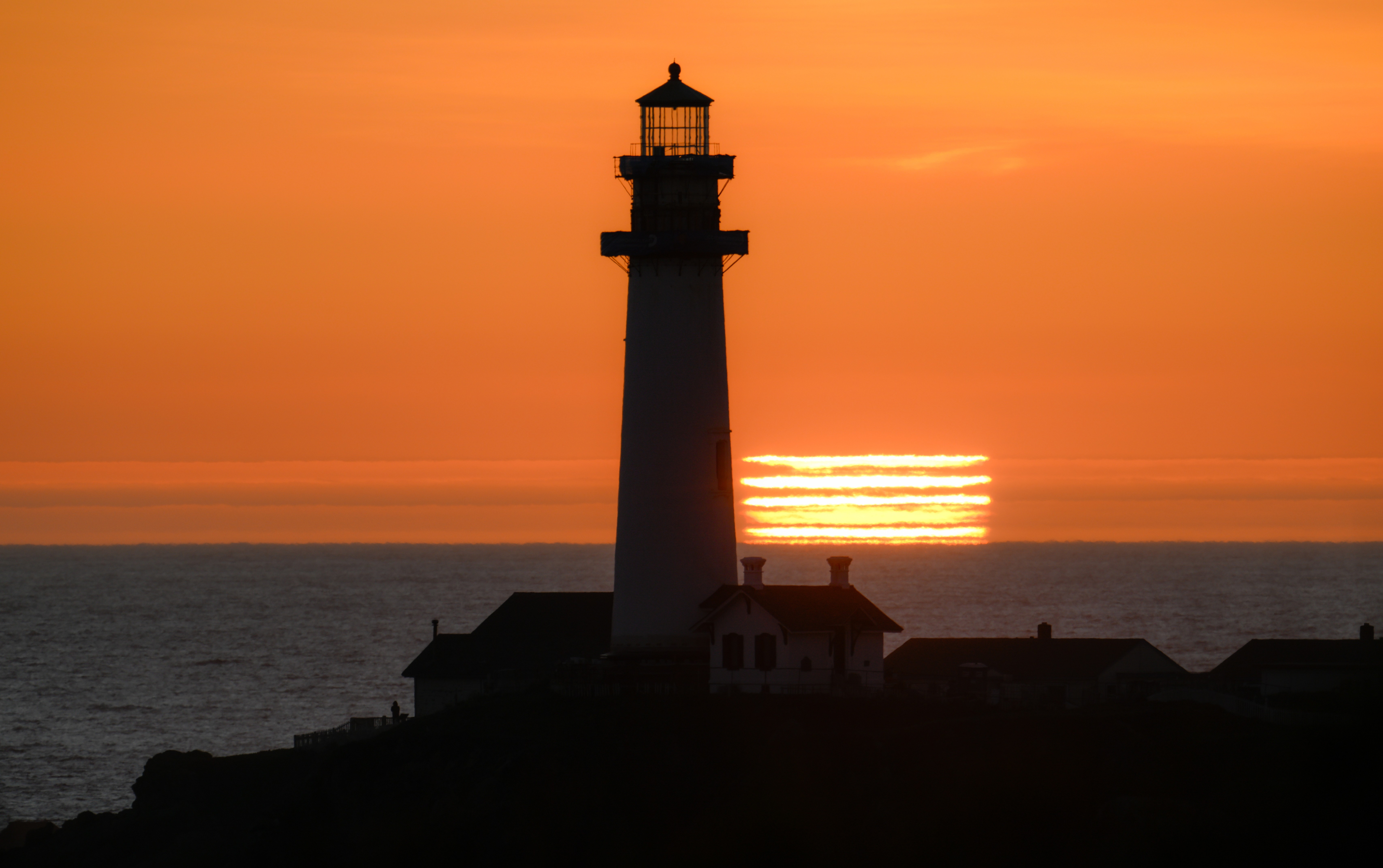 The sun sets behind a lighthouse, with clouds and refraction separating the sun into  about six stripes on the horizon.