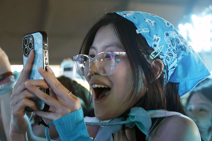 A concertgoer wearing a bright-blue bandanna and clear-framed glasses smiles while taking pictures with their phone.