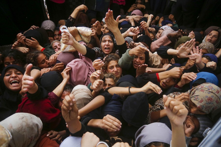 A crowd of people crushes outside a bakery, reaching out to try to buy bread.
