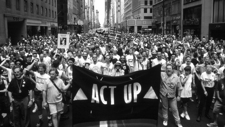 A big group of protesters march down a street holding an ACT UP banner.