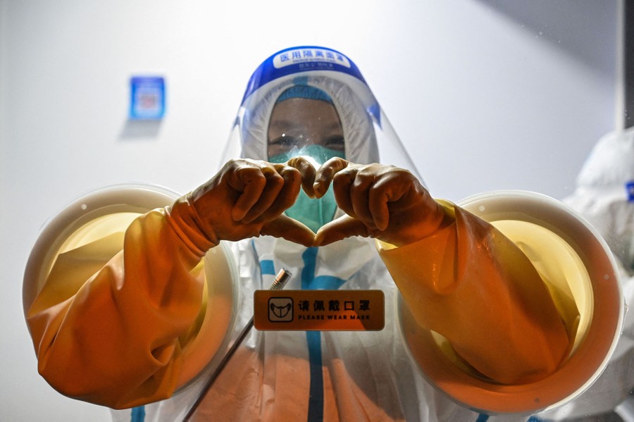 A health worker stands in a booth with her arms poking out through rubber gloves, making a heart shape with her hands.