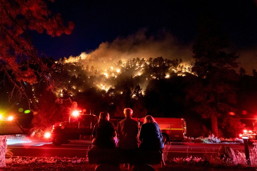 Several people sit alongside a road at night, looking up at a forested hillside on fire.
