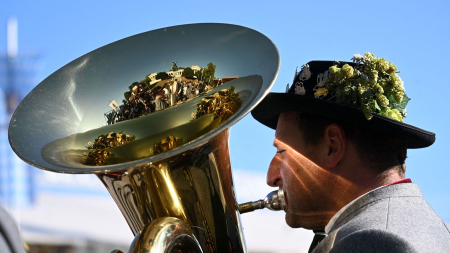 A musician plays a brass instrument outside, wearing a traditional hat.