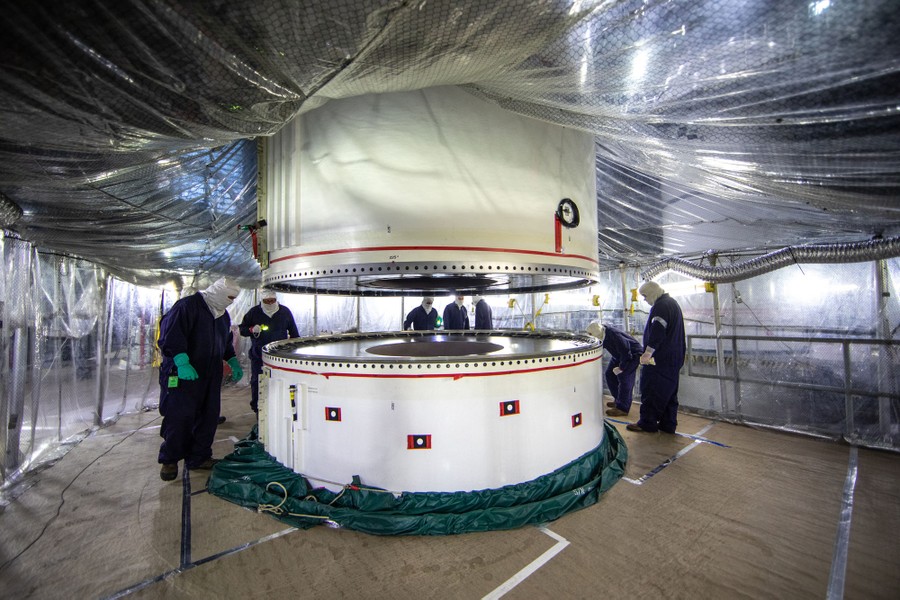 A rocket booster segment is lowered onto another, as about eight workers monitor it closely.