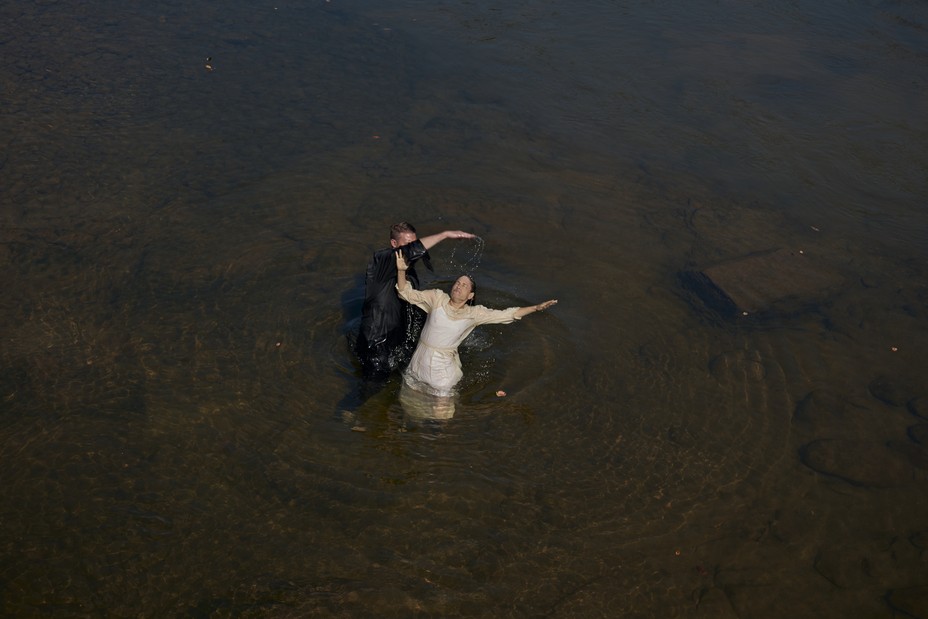 A woman wearing white is baptized in a river by a man in a black gown.