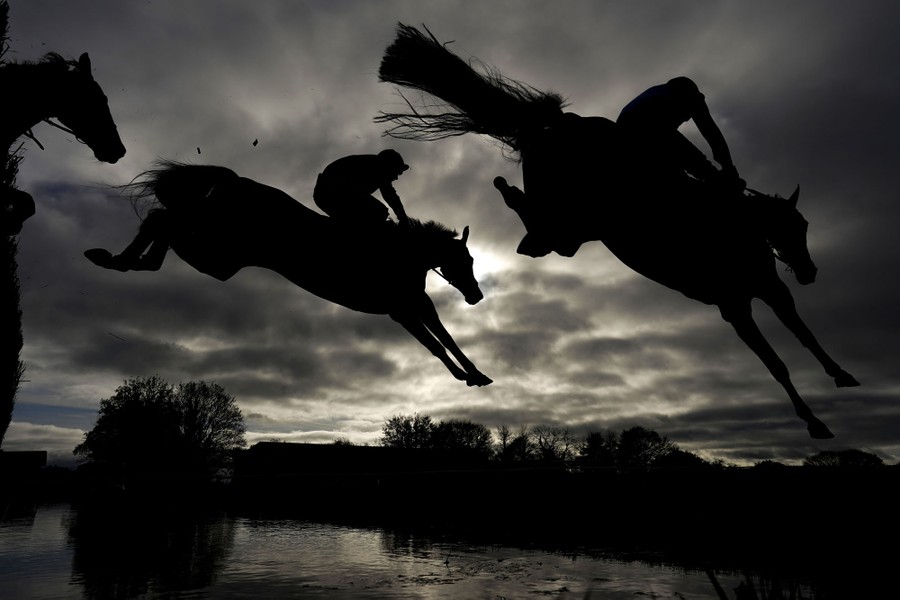 The silhouettes of several leaping racehorses are seen against a cloudy sky.