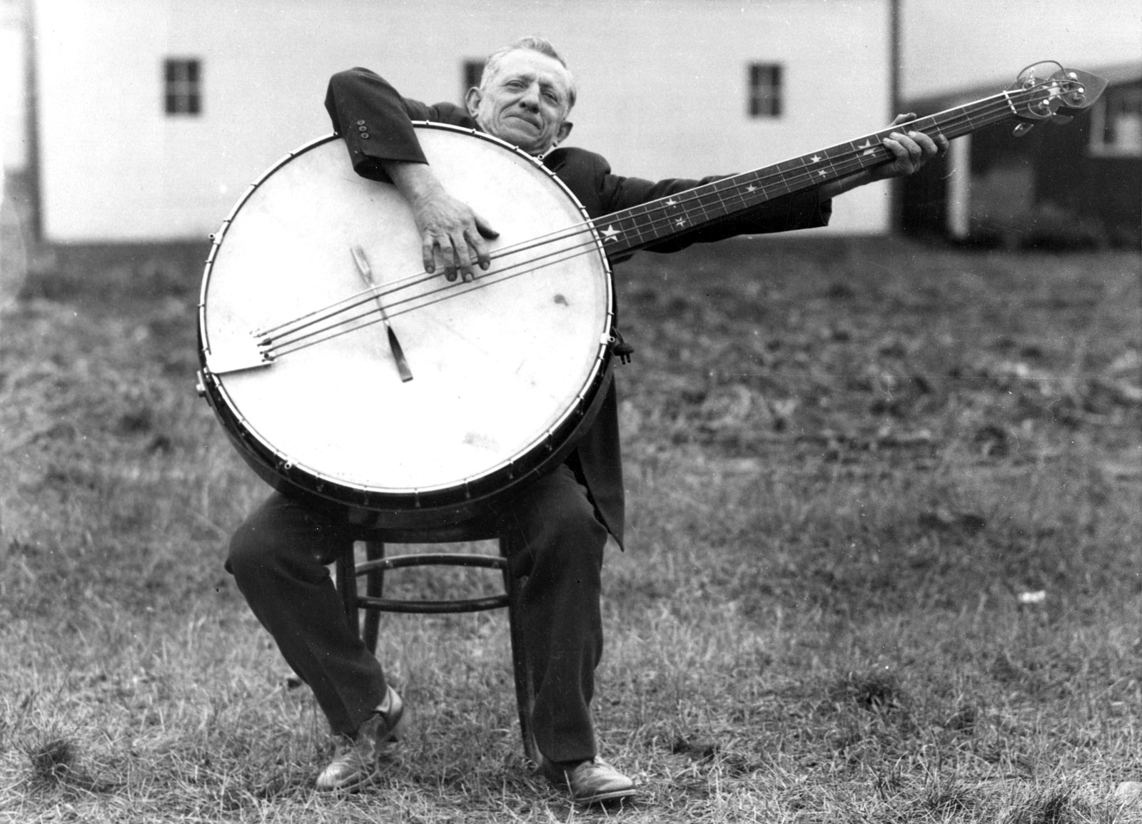 A person sitting on a stool outside plays a gigantic banjo.