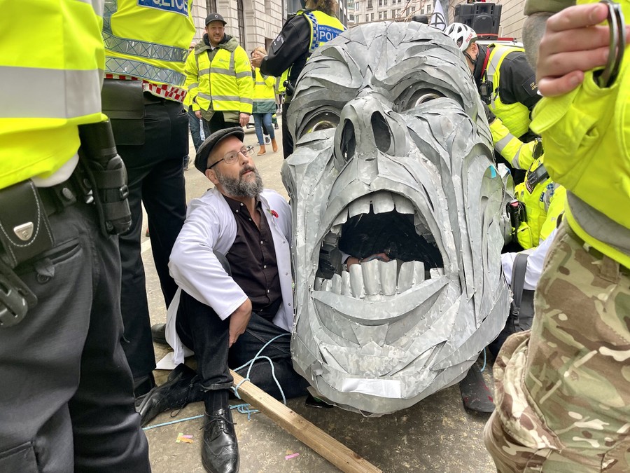 Police officers stand around a protester who sits beside a large sculpture of a human head.