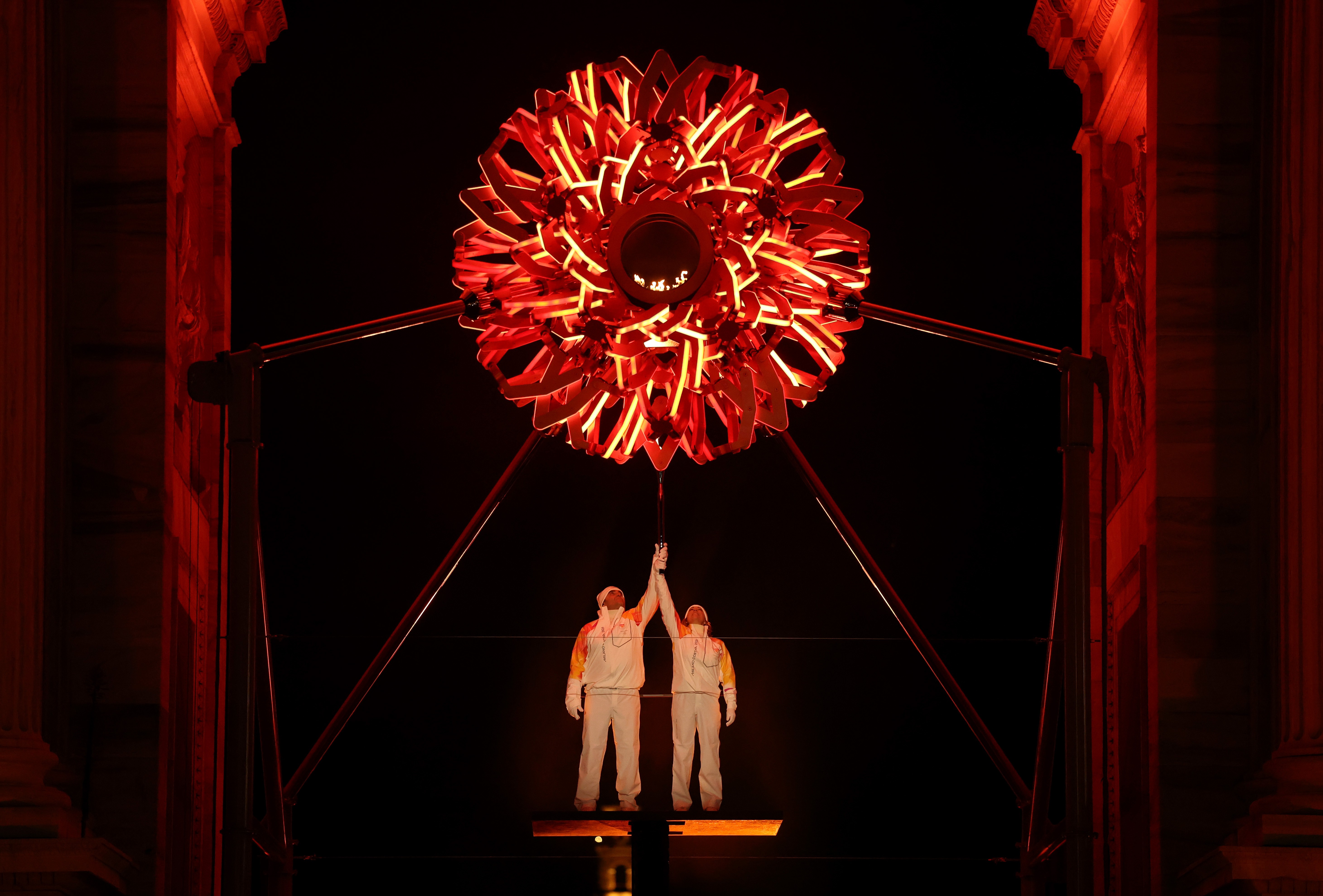 Two athletes jointly hold a torch aloft, standing beneath a tall arch, lighting the ornate spherical Olympic cauldron above them.