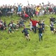 A crowd of spectators stands at the top of a steep hill, watching a couple dozen racers sprint down the hillside.