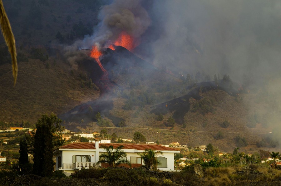 A volcano erupts uphill from a small group of houses.