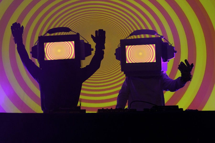 Two performers appear on a stage wearing giant helmets shaped like television sets.