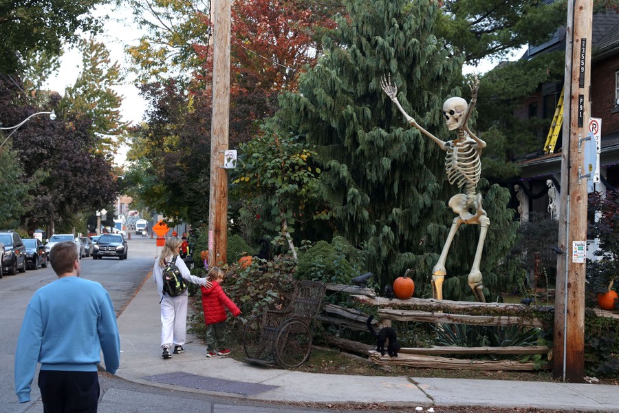 People walk past the yard of a house with a very tall skeleton and other decorations