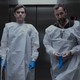 A photo of Patrick Ball and Noah Wyle standing in the elevator with medical gloves on