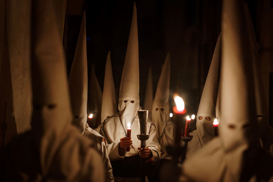 People wearing white costumes with tall conical hats and masks carry candles in a procession.