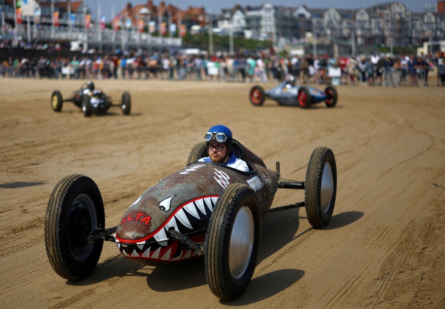 People drive classic race cars on a beach track.