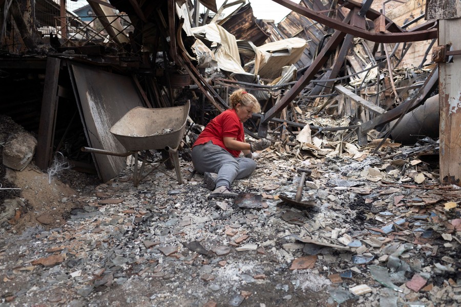 A woman sits amid the debris of a burned house, beside a shovel and a wheelbarrow.