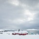 A winter landscape in Greenland; a dark-red house surrounded by snow stands in front of a body of water