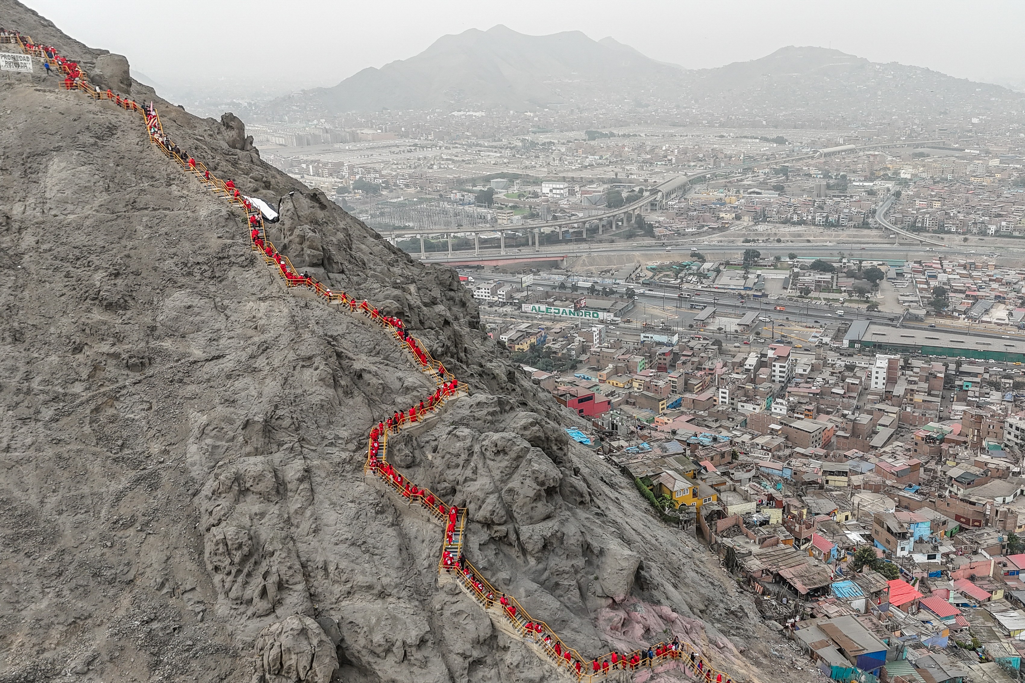 A long line of people attending a memorial are seen standing on a winding path on the side of a rocky mountain.