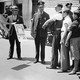 In a black and white photo, health officers at a checkpoint question people in line waiting to be screened to prevent spread of smallpox