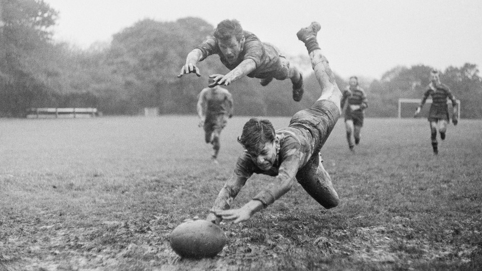 A black and white photograph of a rugby game in action, two players diving for the ball and three running behind them in striped uniforms in the distance on the field
