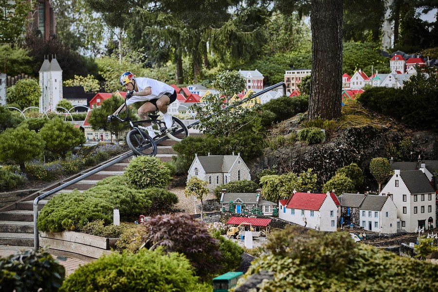 A person rides a bicycle, sliding down a handrail beside steps, among miniature houses in a small park.