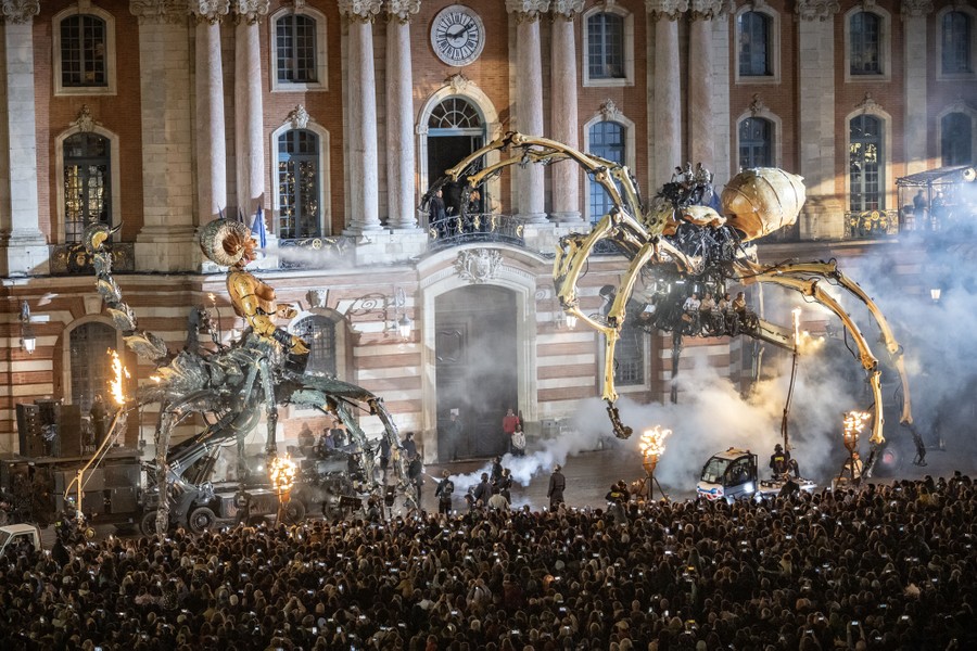 Spectators watch as two giant puppets interact in front of a building.