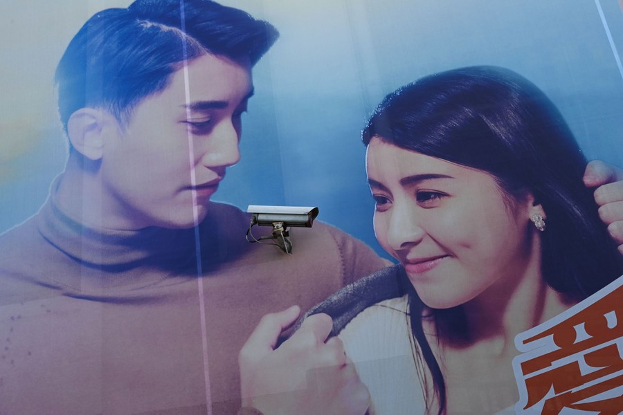 A security camera is situated on a wall, between a man and a woman depicted in a large advertisement mural.