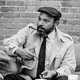 A black-and-white photo August Wilson in a trench coat, driving cap, and tie sitting on sofa and gesturing in conversation with a brick wall behind