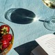 A bowl of strawberries, a glass of wine, and a book open on a tablecloth