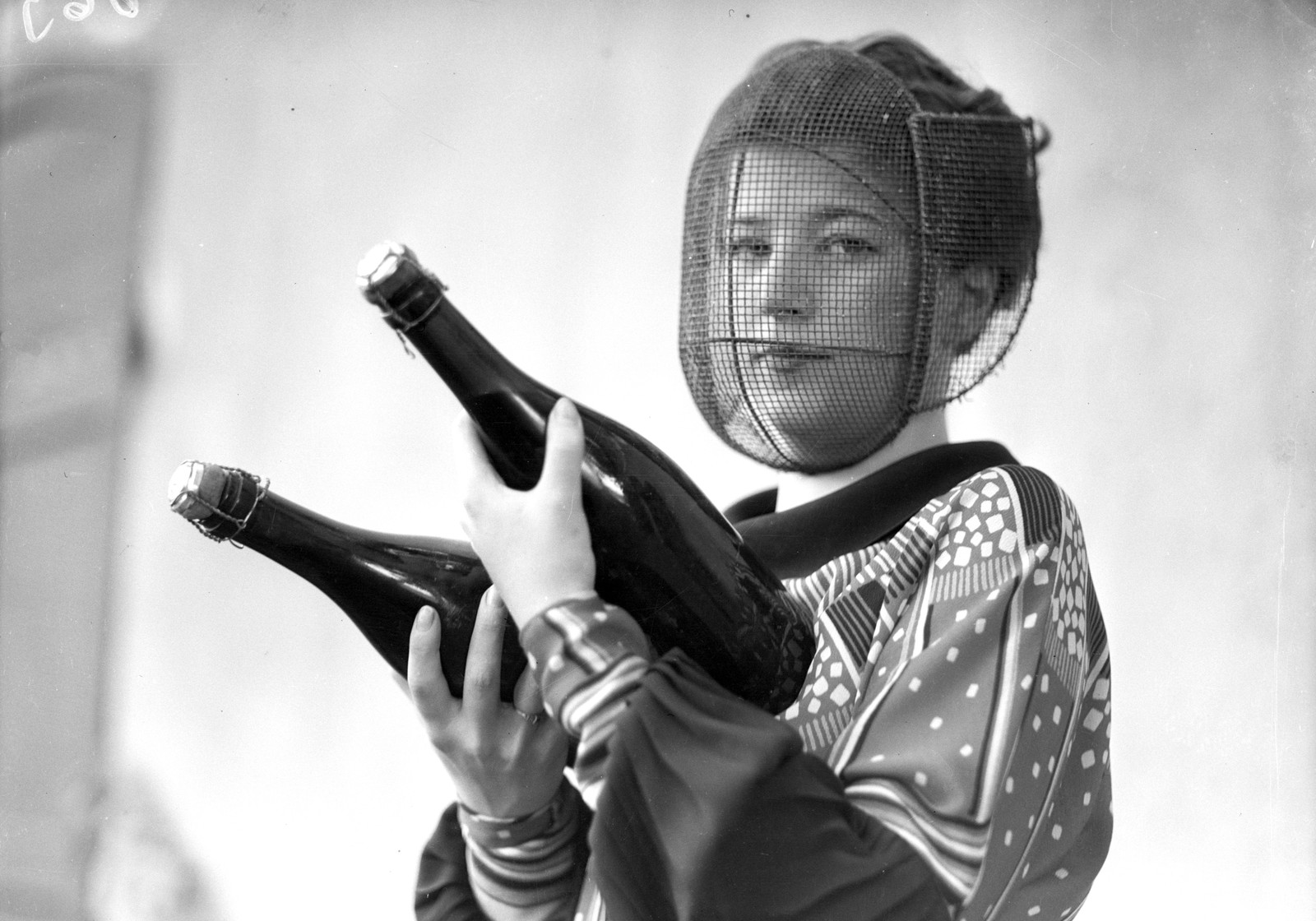 A person holds two champagne bottles in their hands, while wearing a protective mesh face mask.