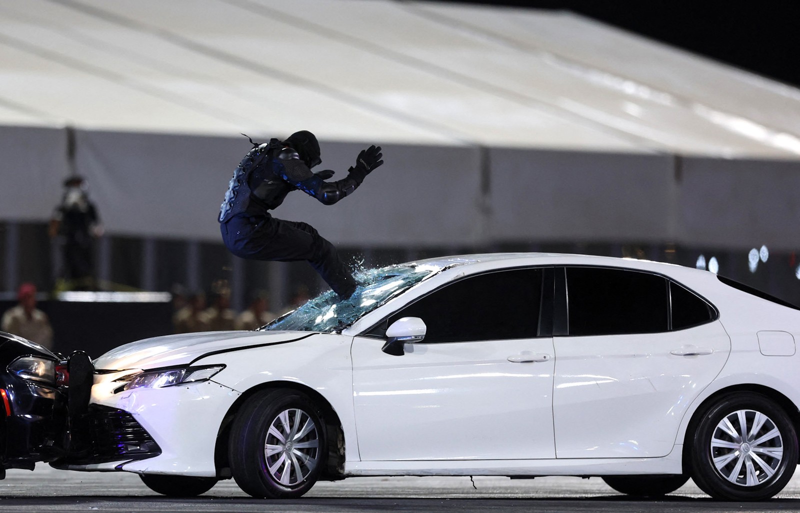 A person wearing protective gear leaps feet-first into the windshield of a moving vehicle during a demonstration.