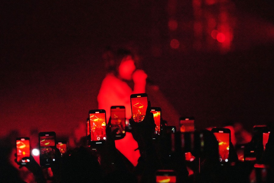 More than a dozen hands hold up mobile devices in the air and record Billie Eilish, who sings in the blurred background.