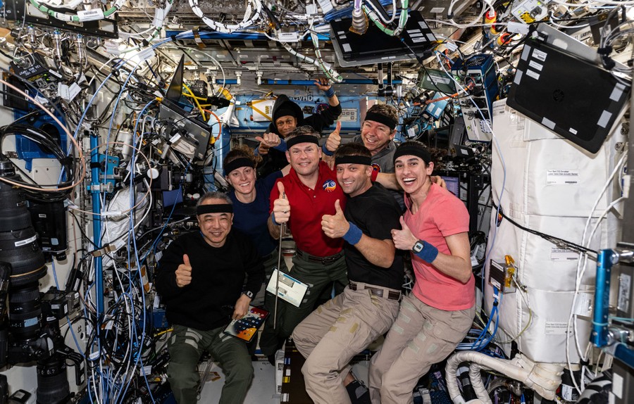 Seven astronauts pose, giving thumbs-up signs while floating inside a cluttered module of the International Space Station.