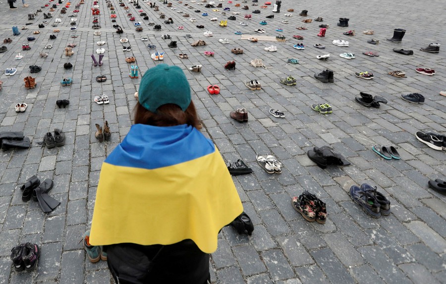 A woman with a Ukrainian flag on her shoulders looks at shoes arranged in a public square as a memorial.