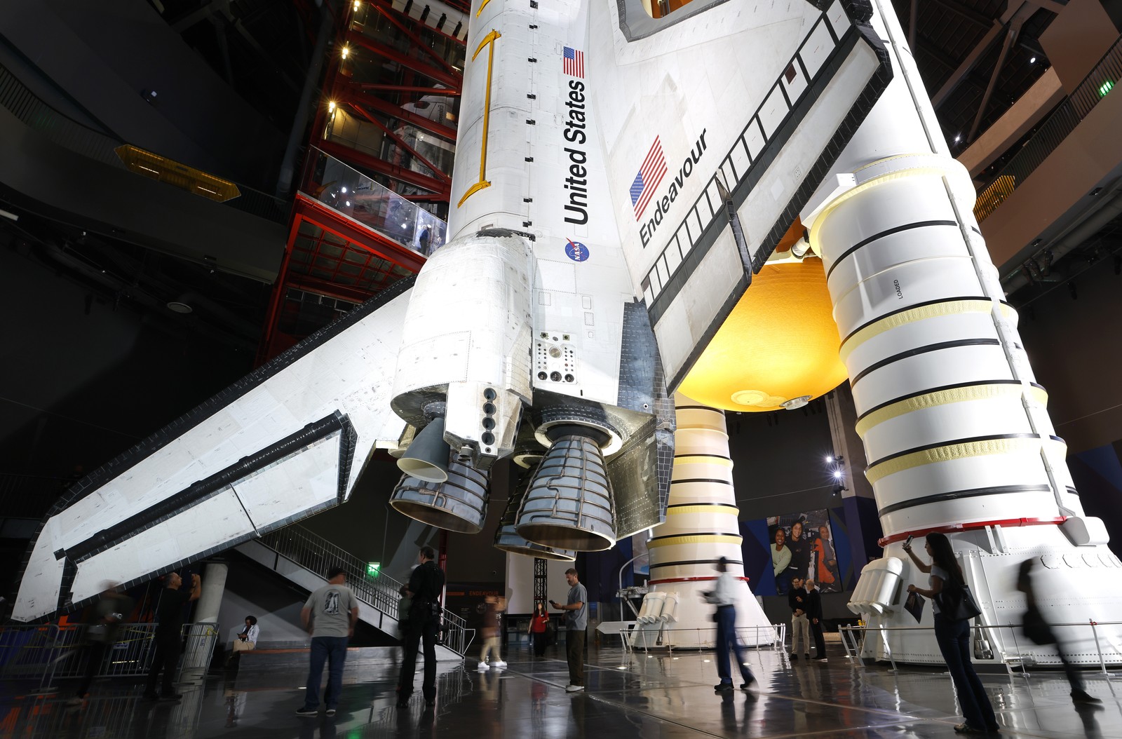 People walk near the base of a decommissioned space shuttle in a museum setting.