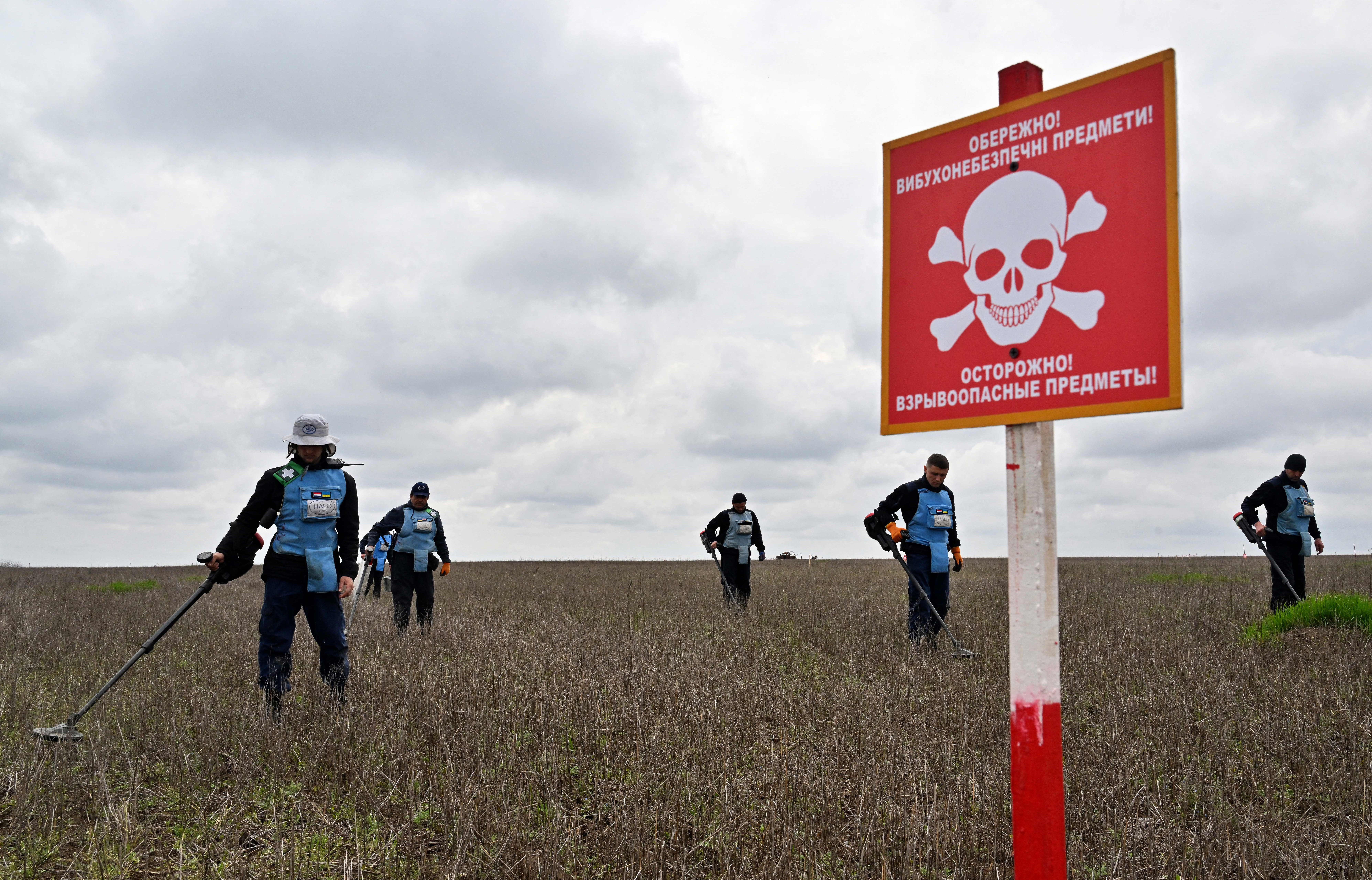 Six people wearing protective gear carry metal detectors while walking through a farm field, near a sign with a skull-and-crossbones warning about demining activity.