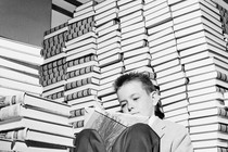 A sitting child reads a book, surrounded by tall stacks of other books