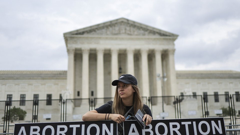 A person in a black cap stands in front of a columned building, behind two signs that each read "Abortion."
